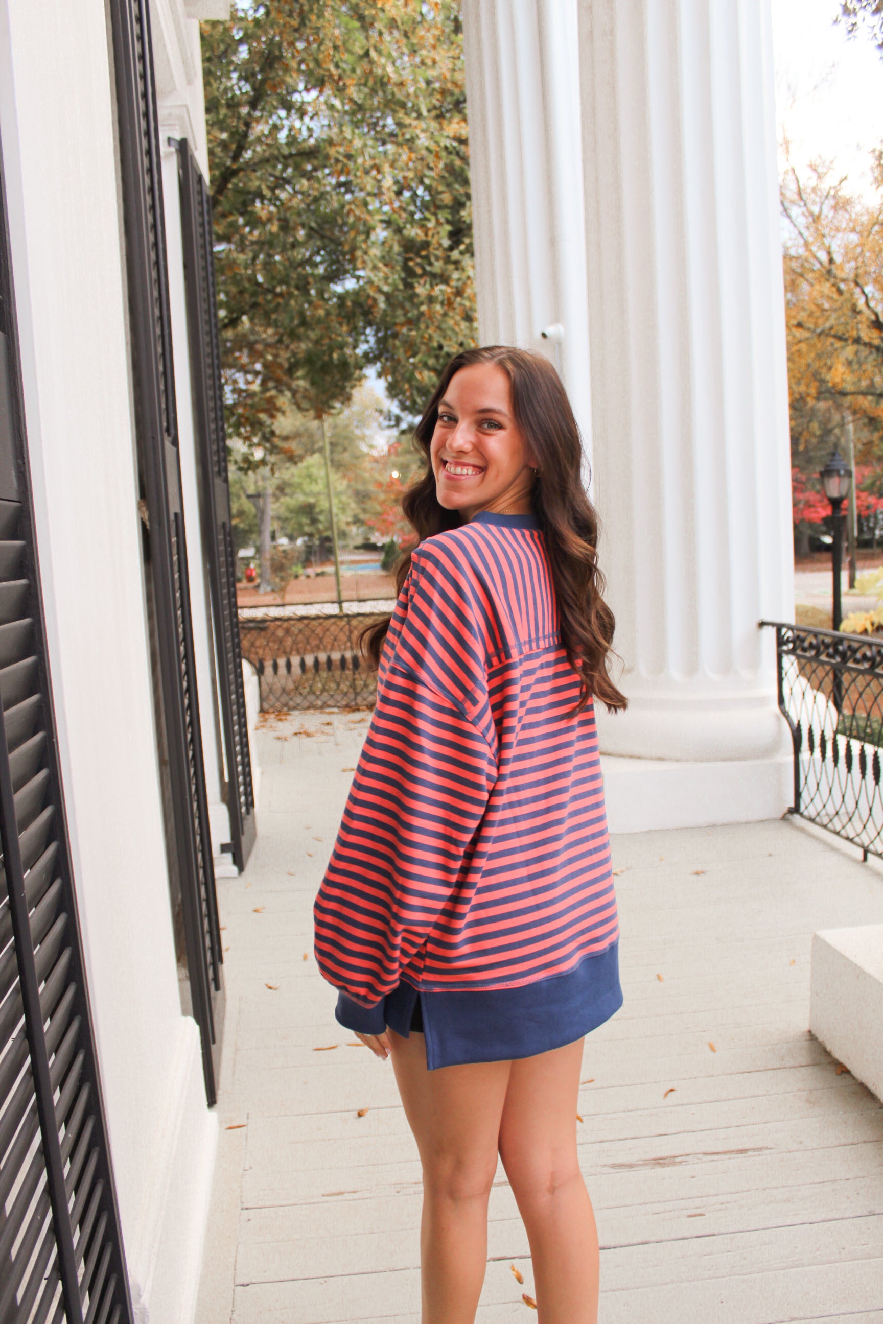 Woman wearing a red and blue striped sweater standing on a porch with columns and trees in the background.